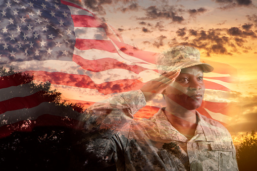 African descent, female USA army soldier overlay on dramatic sunset sky and waving American flag.   She salutes the American flag and wears an army uniform.