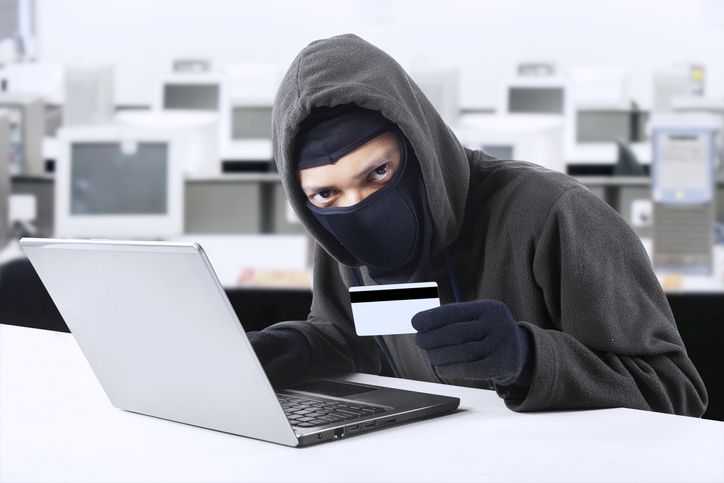 Internet Theft - a man wearing a balaclava and holding a credit card while sat behind a laptop,